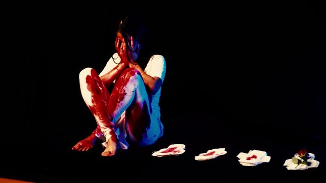 Woman wearing white clothes and being covered in blood, sitting next to 3 sanitary pads also covered in blood and a red rose.