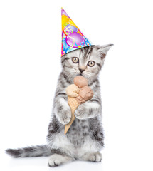 Cute kitten in birthday hat standing on hind legs and holds ice cream. isolated on white background