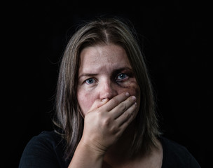 Young beaten up woman looking at camera and covered her mouth with her hands. Isolated on dark background