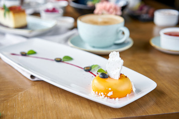 Yellow mousse-cake. Breakfast in the cafe, morning coffee. Cappuccino and lots of desserts on the table. French dessert, blueberry, mint and a piece of meringue on a white plate. Restaurant menu