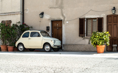 Small vintage italian car. Beige color old car in front of old house facade and flowers..