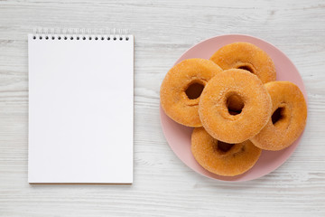 Homemade apple donuts on a pink plate, blank notepad on a white wooden surface, view from above. Top view, flat lay, overhead. Copy space.
