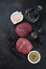 Naklejka premium High angle view of raw marbled beefsteaks on a dark brown stone background with charcoals and decorative grill, vertical shot