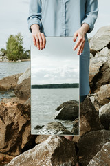 Closeup of woman standind on a shore in blue dress holding a mirror with sea water reflecting in it
