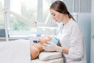 Beautiful blue-eyed girl at the reception of a beautician health clinic. Dermatologist examines the skin under the lamp side view