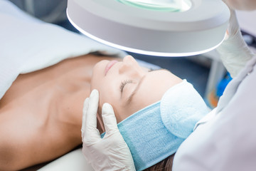 Beautiful blue-eyed girl at the reception of a beautician health clinic. Dermatologist examines the skin under the lamp