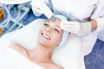 Beautiful blue-eyed girl at the reception of a beautician health clinic. Dermatologist making the cleaning of the skin. Girl watching the beauty and youth