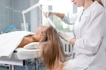 Beautiful girl at the reception of a dermatologist in the health clinic. Dermatologist conducting hair restoration procedure doing hair massage with liquid nitrogen side view
