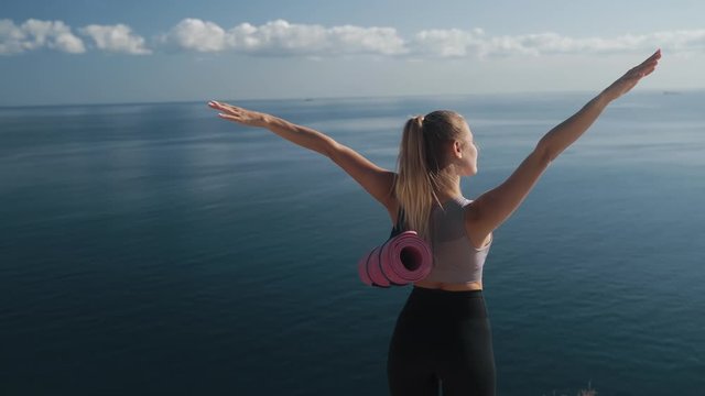 Backside view, woman in sportswear stands to edge of cliff, opens her arms wide