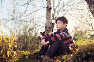 The boy sits on the grass with fallen leaves and looks into the distance.