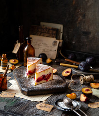 two slices of baked cheesecake or cottage cheese casserole on shortcrust with plums and powdered sugar stands on wooden board on rustic table with plums, old bottles opposite concrete wall