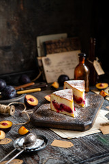 two slices of baked cheesecake or cottage cheese casserole on shortcrust with plums and powdered sugar stands on wooden board on rustic table with plums, old bottles opposite concrete wall