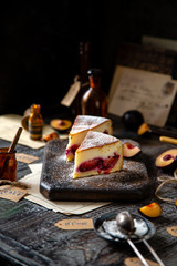 two slices of baked cheesecake or cottage cheese casserole on shortcrust with plums and powdered sugar stands on wooden board on rustic table with plums, old bottles opposite concrete wall