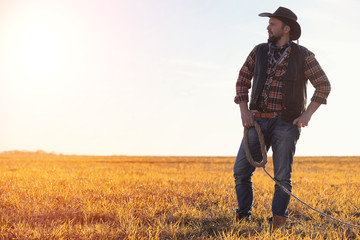 A man cowboy hat and a loso in the field. American farmer in a field wearing a jeans hat and with a loso. A man is walking across the field in a hat