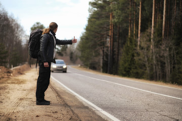 A young man is hitchhiking around the country. The man is trying to catch a passing car for traveling. The man with the backpack went hitchhiking to the south.