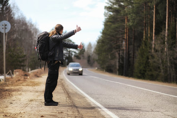 A young man is hitchhiking around the country. The man is trying to catch a passing car for traveling. The man with the backpack went hitchhiking to the south.