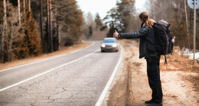 A young man is hitchhiking around the country. The man is trying to catch a passing car for traveling. The man with the backpack went hitchhiking to the south.