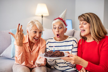 Senior woman blow on birthday cake during a birthday party. Group of seniors celebrating a birthday at home. Senior Woman Blows Out Birthday Cake Candles with females friends.