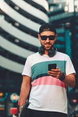 Young man with headphones around neck wearing hat and sunglasses holding a skateboard in his hand...