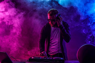 Young stylish man in glasses posing behind mixing console on colored smoke studio background.