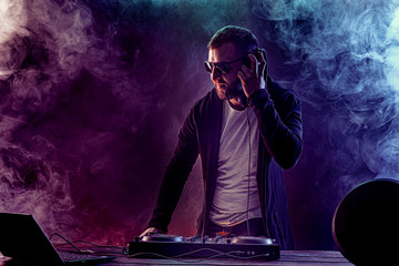 Young stylish man in glasses posing behind mixing console on colored smoke studio background.