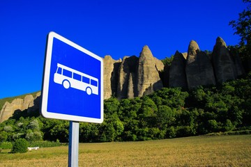 View on isolated bus stop sign against rural field, trees, needle like sharp bizarr rock formations...