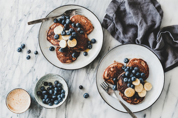 Flat-lay of banana pancakes with fresh blueberry and honey and cup of coffee over white marble table background, top view. Healthy comfort breakfast set
