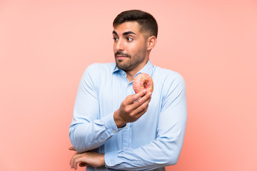 Young man holding a donut over isolated pink background making doubts gesture while lifting the shoulders