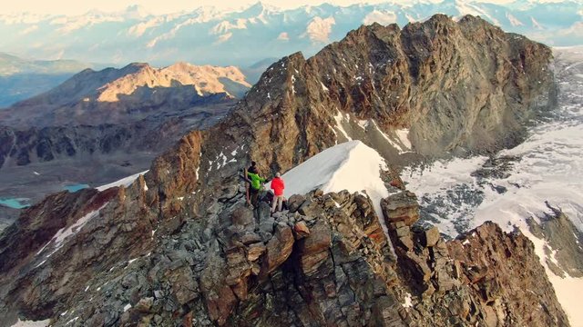 Stunning panoramic aerial view of three hikers standing on the peak of Lagginhorn mountain (4010 metres above sea level) in the Switzerland alps just after sunrise. Drone orbiting around the summit.