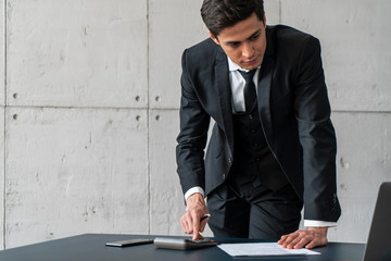 Serious young caucasian businessman in dark suit using calculator and laptop in loft office....