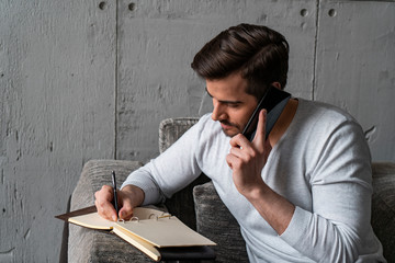Portrait of handsome bearded businessman in casual clothes talking on smartphone and writing in...
