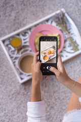Young woman food blogger takes blog photo of coffee latte, orange juice and avocado fresh toasts. Closeup of women's hands making photo of healthy breakfast on a tray with mobile phone. vertical photo