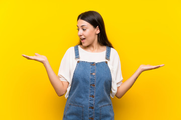 Young woman in dungarees over isolated yellow background holding copyspace with two hands