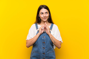 Young woman in dungarees over isolated yellow background scheming something