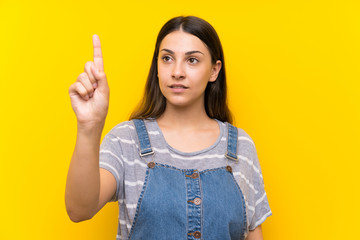 Young woman in dungarees over isolated yellow background touching on transparent screen