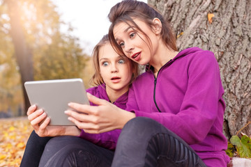 Outdoors leisure. Sisters sitting near tree in the autumn park with digital tablet watching video shocked close-up
