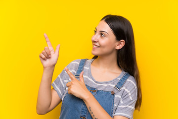 Young woman in dungarees over isolated yellow background pointing with the index finger a great idea