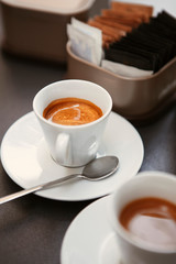 Close up. The small white ceramic mug (cup) with Italian espresso (coffee) on a table with a saucer and a metal spoon in a local Italian bar (coffee shop) in Milan, Lombardy, Italy. Beverage.
