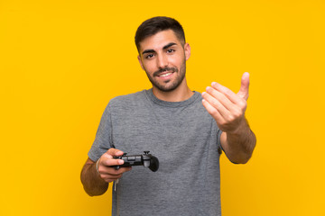 Young handsome man playing with a video game controller over isolated yellow background inviting to come with hand. Happy that you came - obrazy, fototapety, plakaty