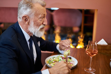Elegant caucasian senior male sits in restaurant. Bearded man look and eat. Fireplace behind him. Horizontal image
