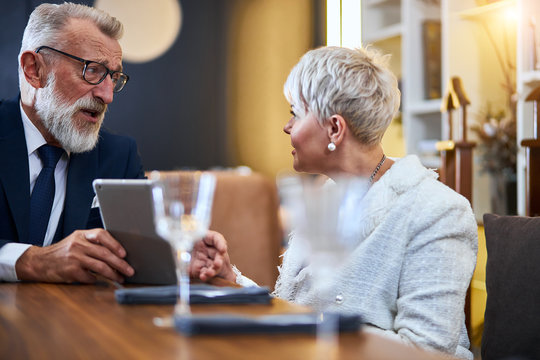 Joyful, cheerful, mature couple in love having time at restaurant while chatting and looking at each other. Senior man show something in tablet