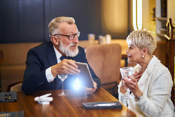 Senior rich couple websurfing on internet with tablet, man in tuxedo, woman in white blazer talking in restaurant