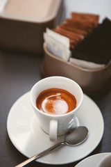 Close up. The small white ceramic mug (cup) with Italian espresso (coffee) on a table with a saucer and a metal spoon in a local Italian bar (coffee shop) in Milan, Lombardy, Italy. Beverage.