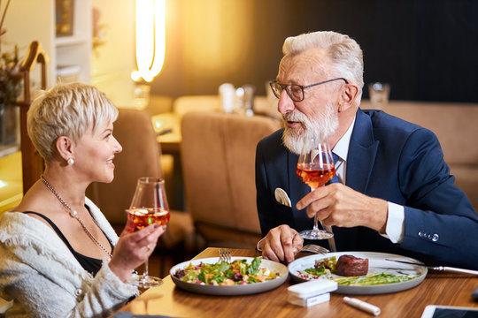 Stylish, elegant senior couple look at each other and chatting, close relationship. Grey-haired man in tuxedo raise a glass. Use e-cigarette, vape. Modern technologies