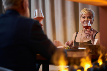 Happy gray-haired elderly lady in black dress toasting with glass of champagne to male in elegant restaurant, look at him. Rear view on man in tuxedo. Romantic setting