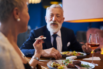 Senior caucasian bearded male sits and look at woman with e-cigarette, iqos in restaurant hotel. Elegant man in tuxedo. Modern technologies concept