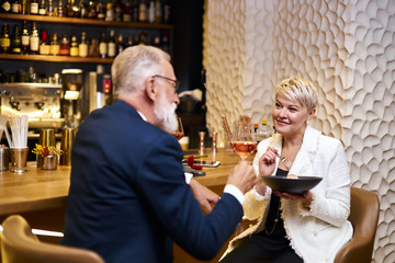 Mature couple of caucasian man and woman sit in restaurant and eat dessert, drink glass of wine. Male in tuxedo, female in white blazer. Woman holding dish