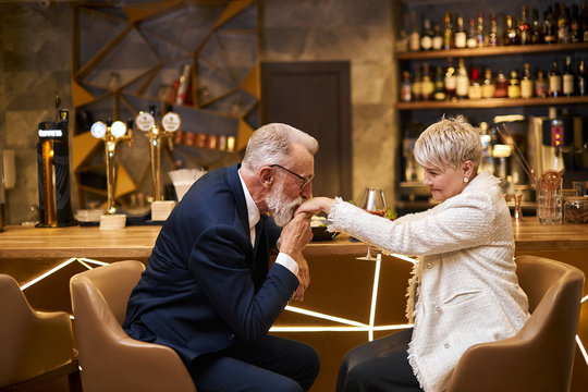 Handsome man in beautiful tuxedo and woman in white blazer sit in expensive beautiful restaurant. Male kissing hand and declare his love. Romantic image, light view