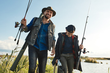 Happy son and father go to fishing place. They hold fishing rods, wear clothes for fishing. Sunny warm day.