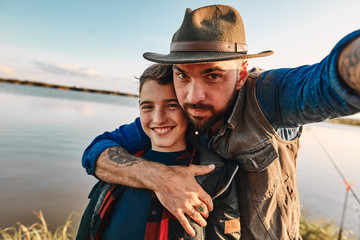 Father hugs son and takes selfie. They have fun. Background lake.